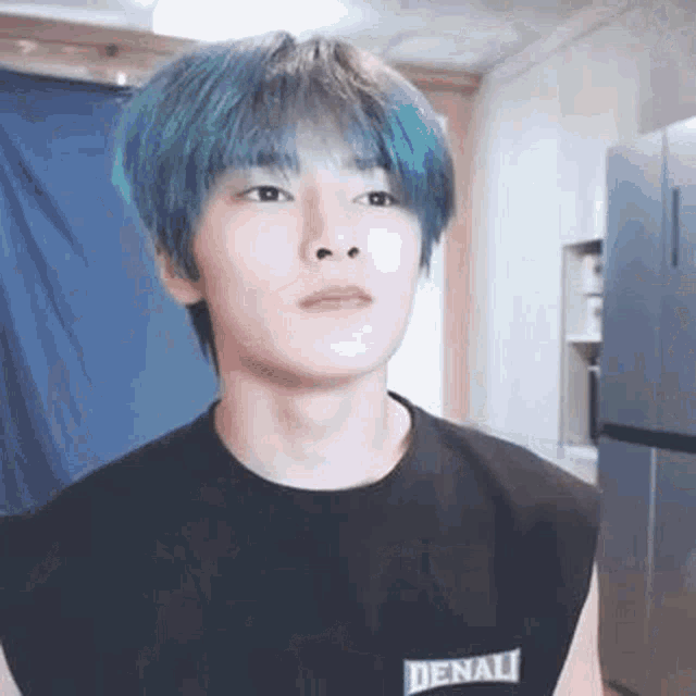 a young man with blue hair and a black shirt is standing in front of a refrigerator in a room .