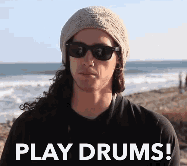 a man wearing sunglasses and a beanie says play drums on the beach