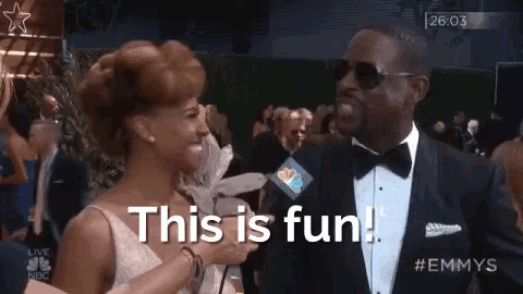 a man in a tuxedo is talking to a woman on a red carpet and the woman is saying this is fun