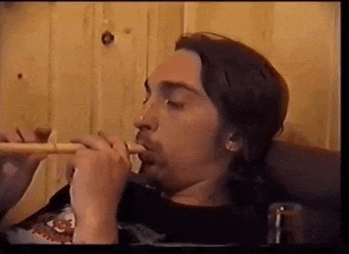 a man with long hair is smoking a pipe in front of a wooden wall .