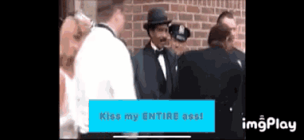 a man in a tuxedo is standing in front of a brick wall with a blue sign that says kiss my entire ass