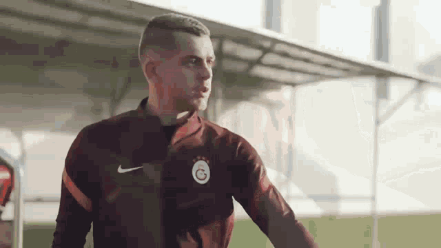 a man in a maroon nike jacket is standing on a soccer field and talking to someone .