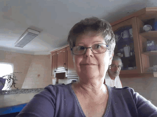 a woman wearing glasses takes a selfie in a kitchen with a man in the background