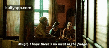 a group of people are sitting around a table in a dark room and one of them is talking about meat in the fridge .