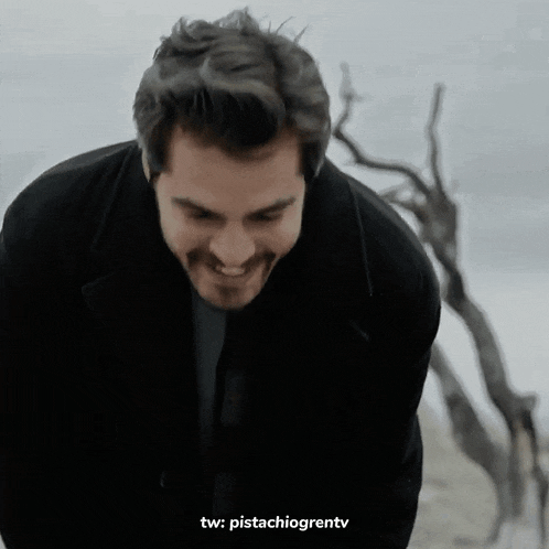 a man in a black jacket smiles in front of a dead tree
