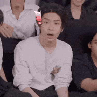 a man in a white shirt is sitting in a group of people and making a funny face .