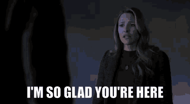 a woman is standing in front of a man in a dark room and saying `` i 'm so glad you 're here ''