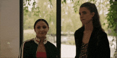 two women are standing next to each other in front of a window and smiling