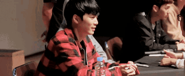 a man in a plaid shirt is sitting at a table with a bottle of water in his hand .