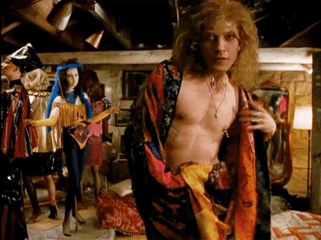 a man without a shirt is standing in a room surrounded by people