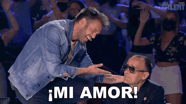 a man in a denim jacket talks to another man in front of a crowd with the words mi amor in white letters