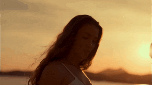 a woman in a white tank top is standing in front of a body of water at sunset .