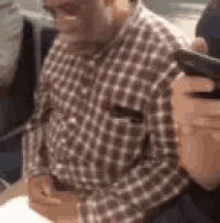 a man in a plaid shirt is sitting at a table with a person holding a cell phone .