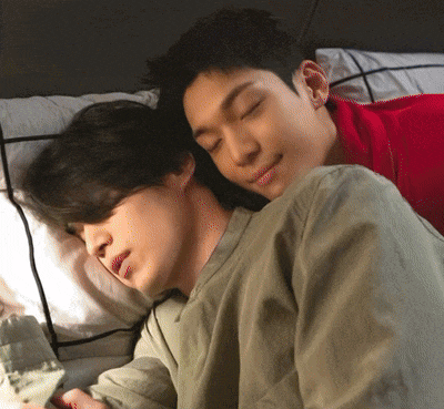 a couple of men laying on a bed with one wearing a red shirt