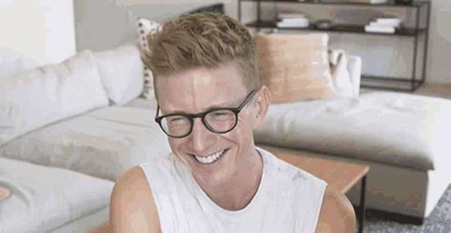 a man wearing glasses and a white tank top smiles in a living room