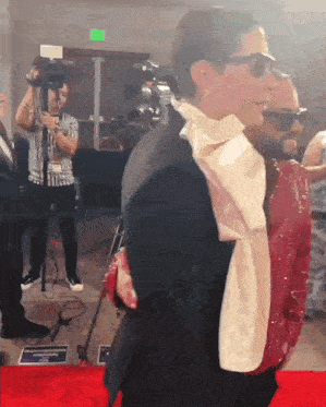 a man in a suit stands on a red carpet with a cameraman behind him
