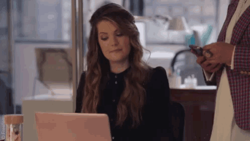 a woman is sitting at a desk using a laptop computer while another woman holds a cell phone .