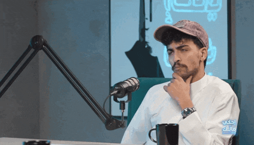 a man wearing a hat is sitting in front of a microphone with arabic writing on the bottom