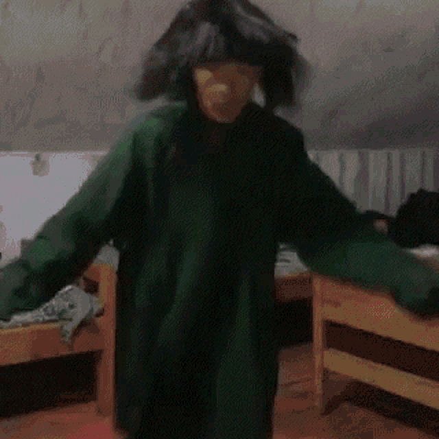 a person wearing a wig and a green shirt is dancing in a room with two beds .