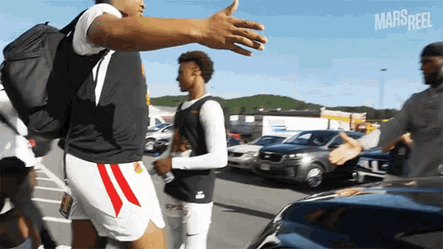 a man with a backpack reaches out to shake another man 's hand in a parking lot with mars reel written on the bottom