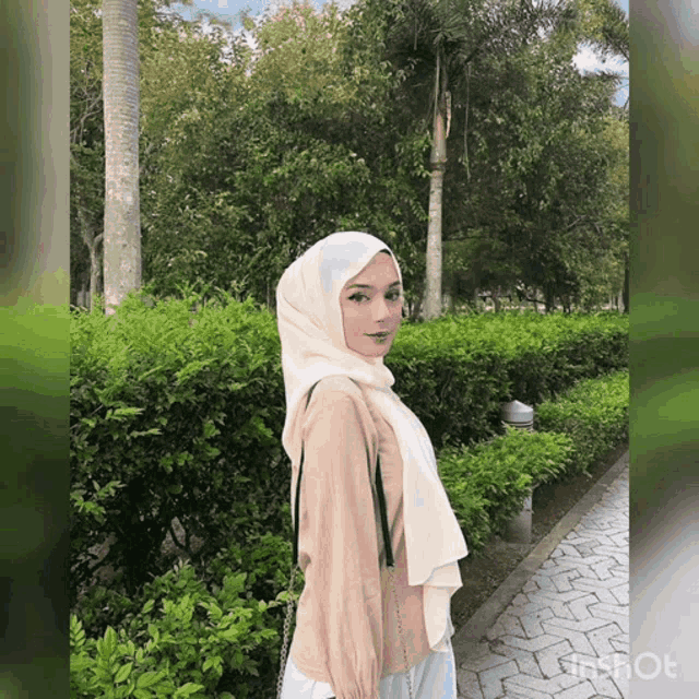 a woman wearing a white hijab stands in front of a bush