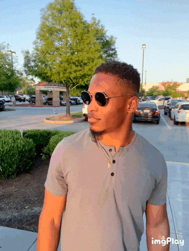 a man wearing sunglasses and a grey shirt is standing in a parking lot with cars parked in the background