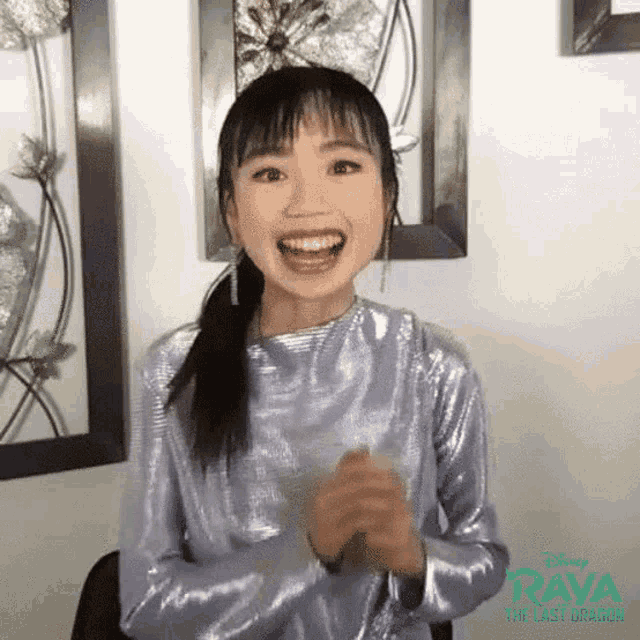 a woman in a silver dress is smiling and clapping her hands in front of a wall with pictures on it .