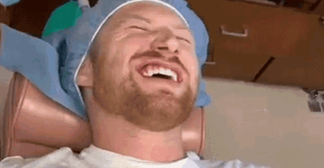 a man with a beard is sitting in a dental chair laughing while a dentist examines his teeth .