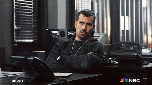 a man sits at a desk with his arms crossed and a nbc logo in the background