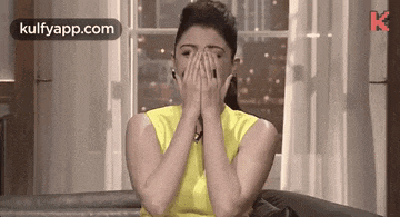 a woman in a yellow shirt is covering her face with her hands while sitting on a couch .