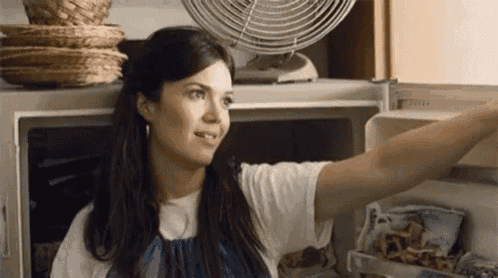 a woman is opening a refrigerator door in a kitchen with a fan in the background .