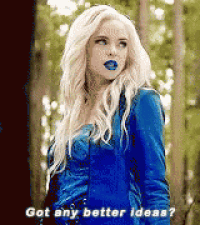a woman with blonde hair and blue lips is wearing a blue jacket and standing in a forest .