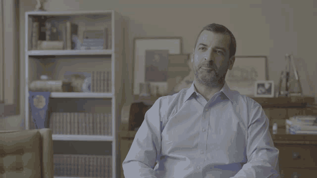 a man in a blue shirt sits in front of a bookshelf with a trophy on it