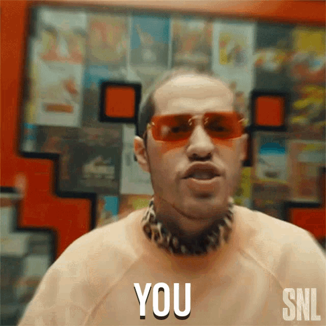 a man wearing sunglasses says " you " in front of a wall of magazines