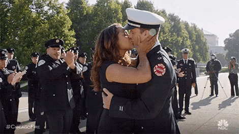 a man and a woman kissing in front of a crowd of firefighters .