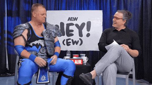 two men sit in front of a sign that says aew hey