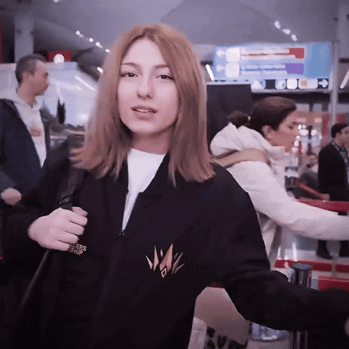 a woman wearing a black jacket with a gold logo on the front is standing in an airport
