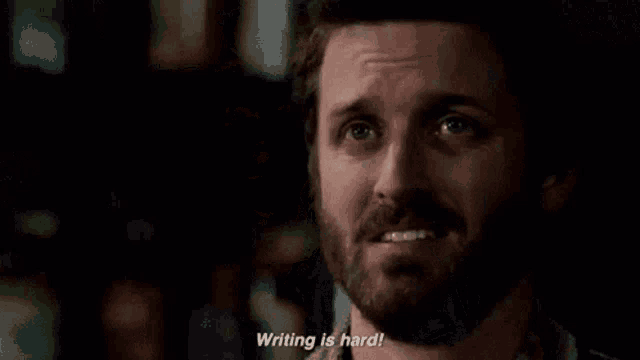 a man in a jacket is standing in front of a window and says writing is hard .