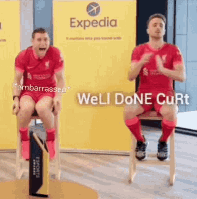 two men in red soccer jerseys are sitting on chairs in front of a yellow sign that says expedia .