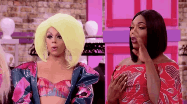 two drag queens are standing next to each other in a room and one of them is wearing a wig .