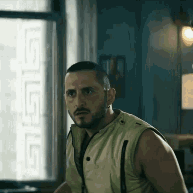 a man with a beard wearing a yellow vest is standing in front of a window in a room .