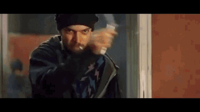 a man in a beanie is smoking a cigarette and pointing at the camera .