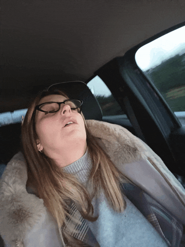 a woman wearing glasses and a fur coat is sleeping in a car