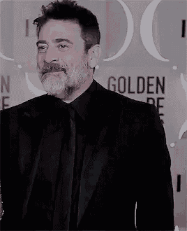a man with a beard is wearing a black suit and tie and smiling .