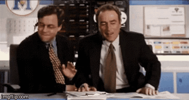 two men in suits and ties are sitting at a desk in front of a sign that says m