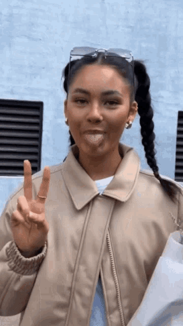 a woman wearing a tan jacket is sticking her tongue out and giving a peace sign