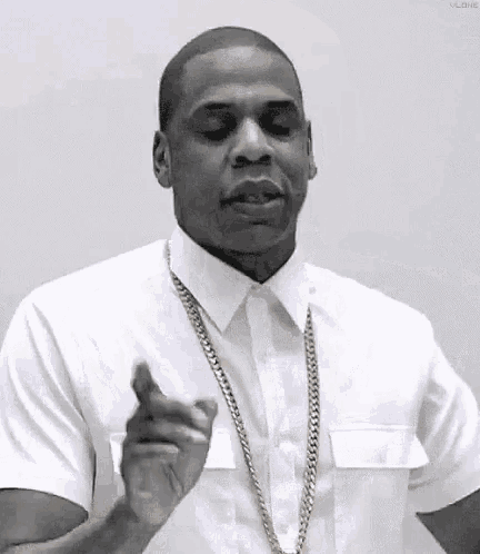 a man wearing a white shirt and a gold chain is pointing at the camera .