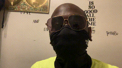 a man wearing sunglasses and a black scarf stands in front of a wall that says " is so good all the me "