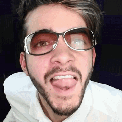 a man with a beard wearing sunglasses and a white shirt is sticking his tongue out