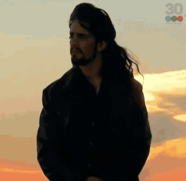 a man with long hair and a beard stands in front of a sunset with the number 30 on the bottom